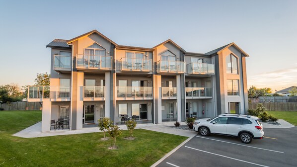 A multi-story residential building with balconies, a parking area, and a small lawn.
