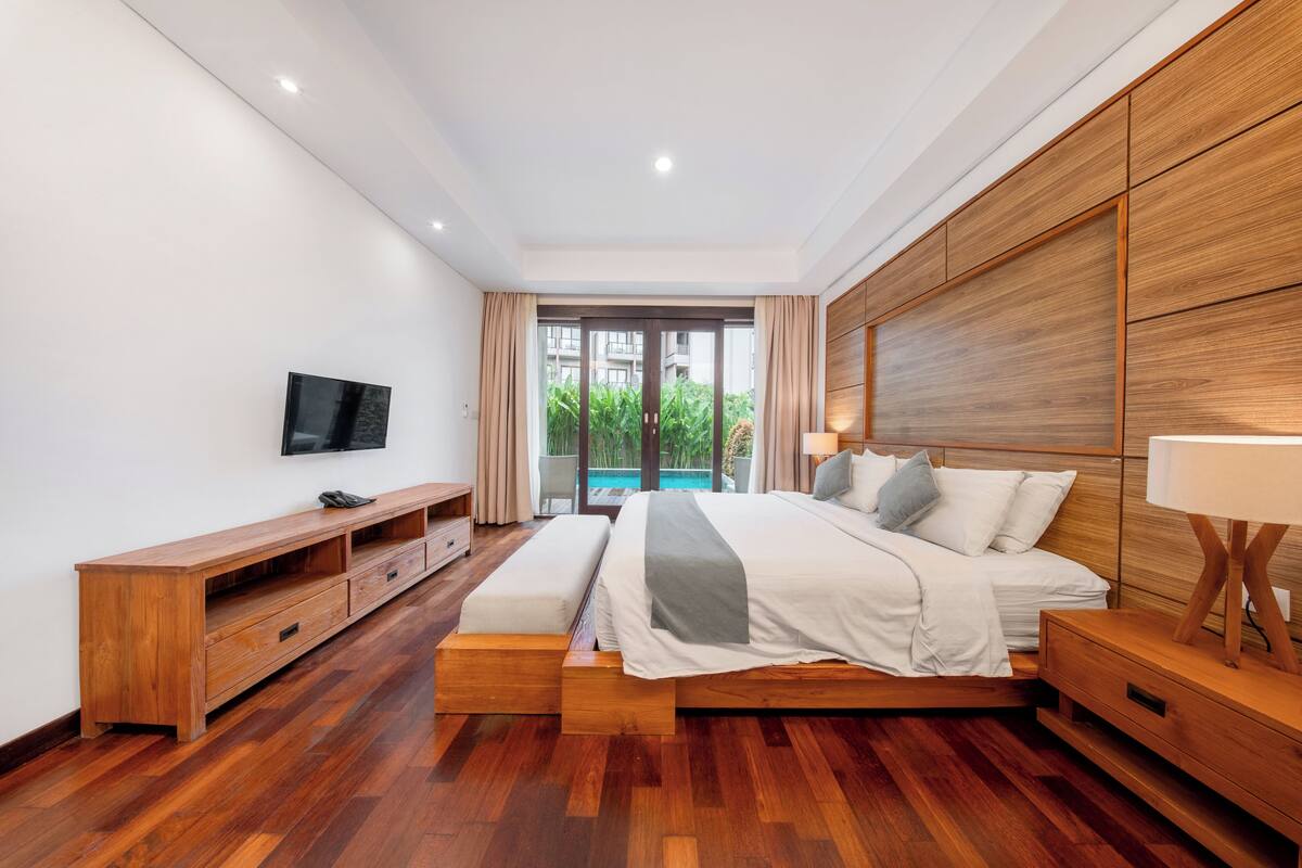 A modern bedroom with a wooden bed, a TV, and a view of a pool.