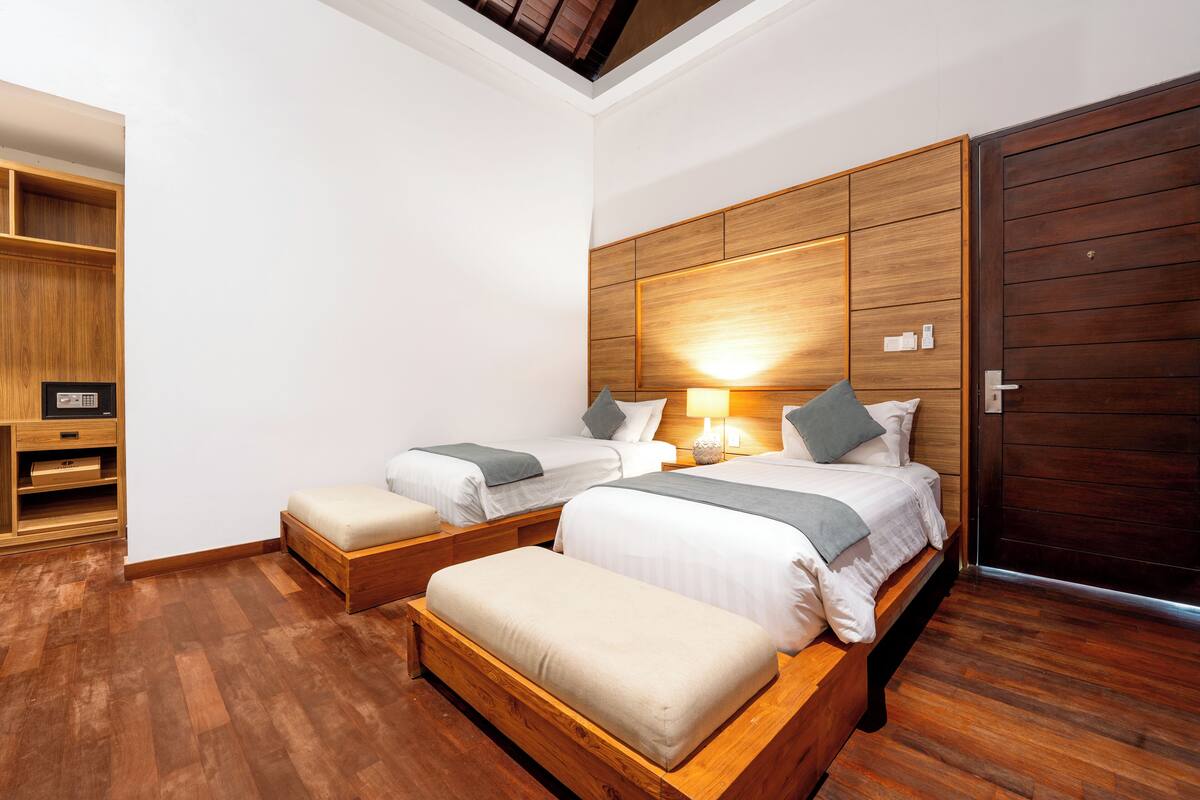 A hotel room with two beds, a wooden bench, a wooden wardrobe, and a ceiling light.
