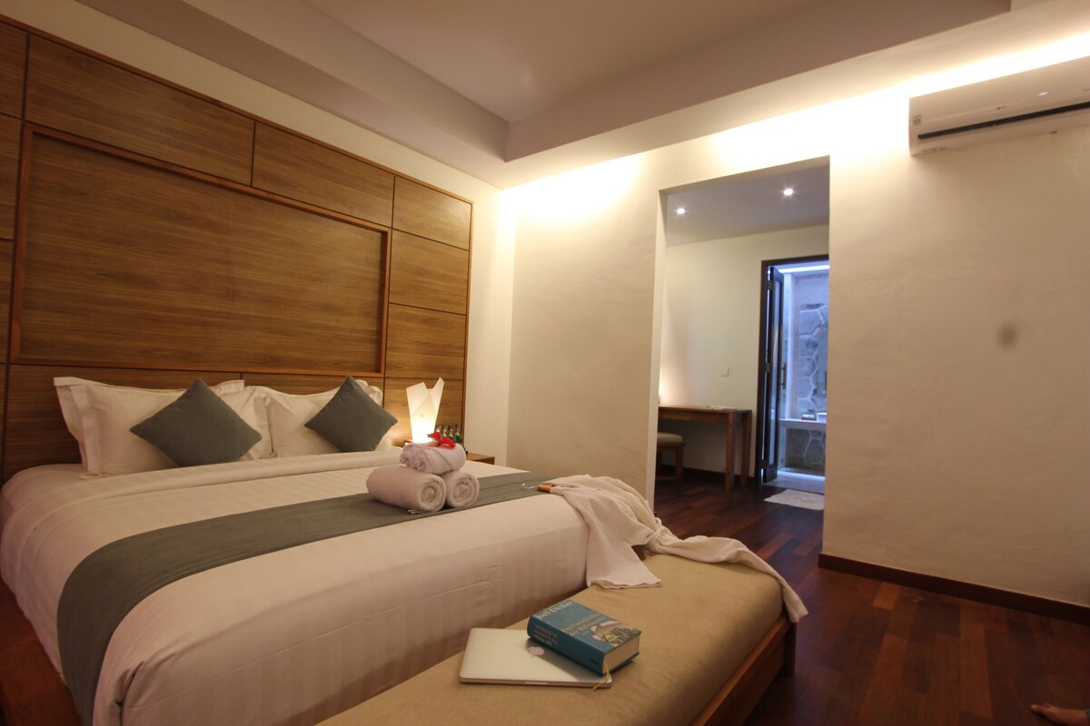 Habitación de hotel con una cama grande, una mesita de noche con lámpara, un cabecero de madera y vistas al baño.