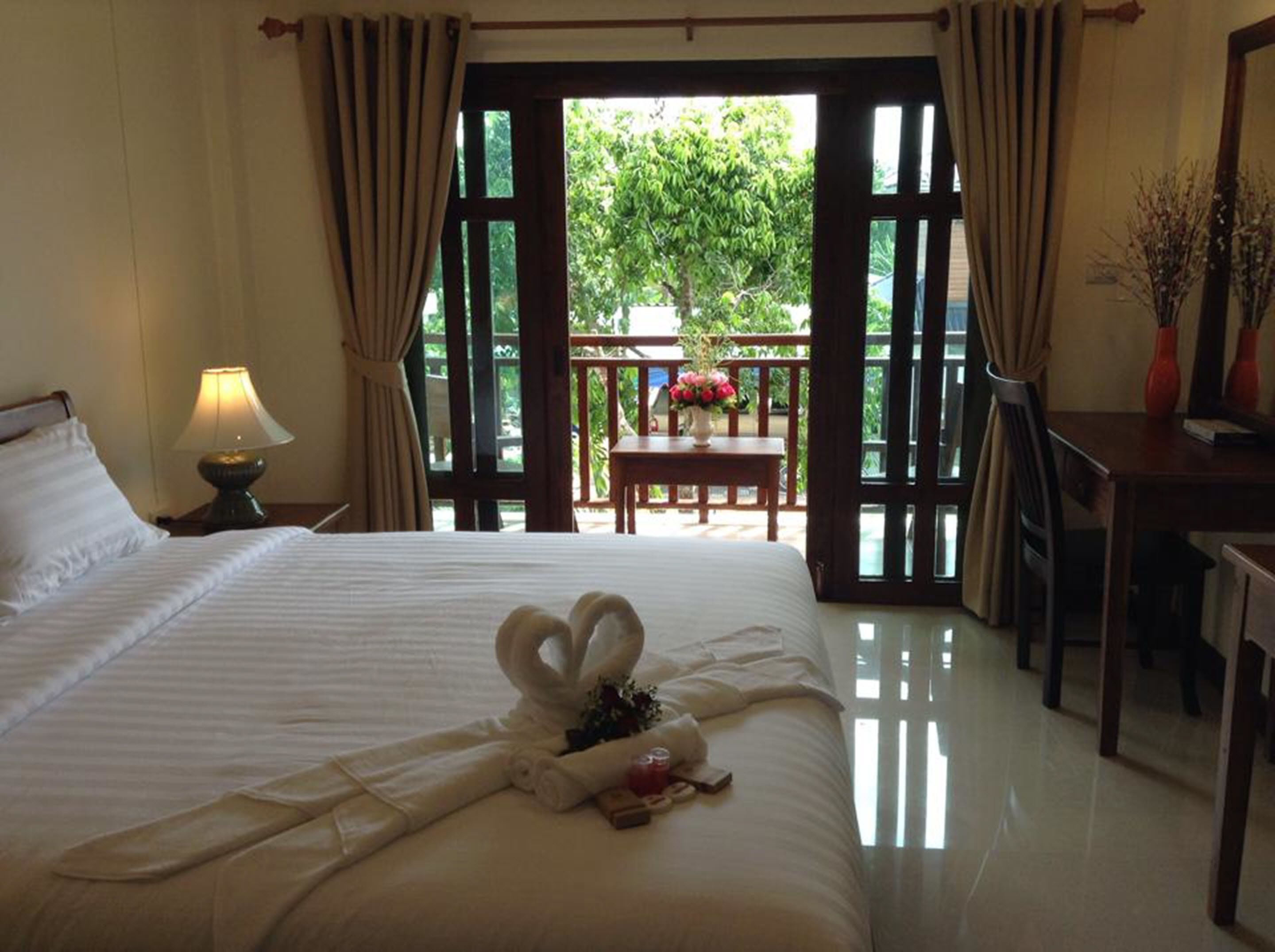A hotel room with a bed, a nightstand with a lamp, a table with flowers, and a view of the outdoors.