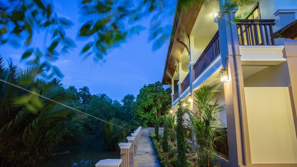 A pathway leading to a building with a balcony, surrounded by lush greenery and a calm water body. - Oh Amphawa Boutique Resort (Amphawa)
