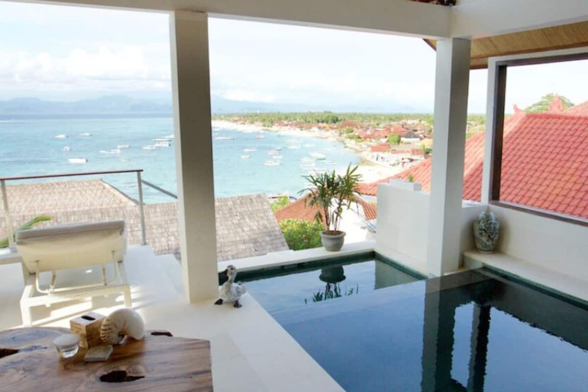 Un balcon avec vue sur la mer et les bâtiments, une piscine et une table ornée d’un vase et d’un chat.