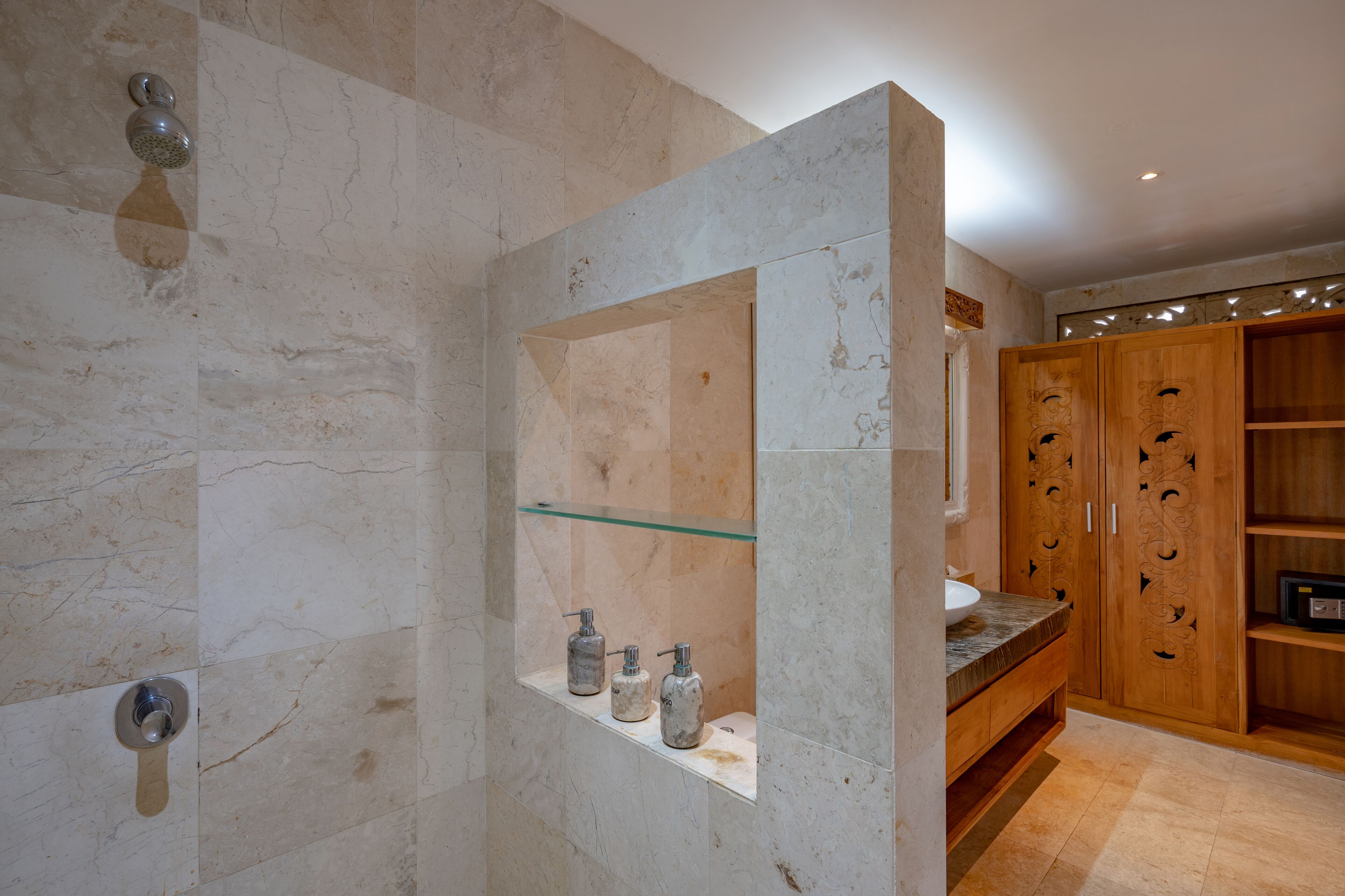 Une salle de bain avec une douche en marbre, une étagère en verre où sont rangés les articles de toilette et des meubles en bois.