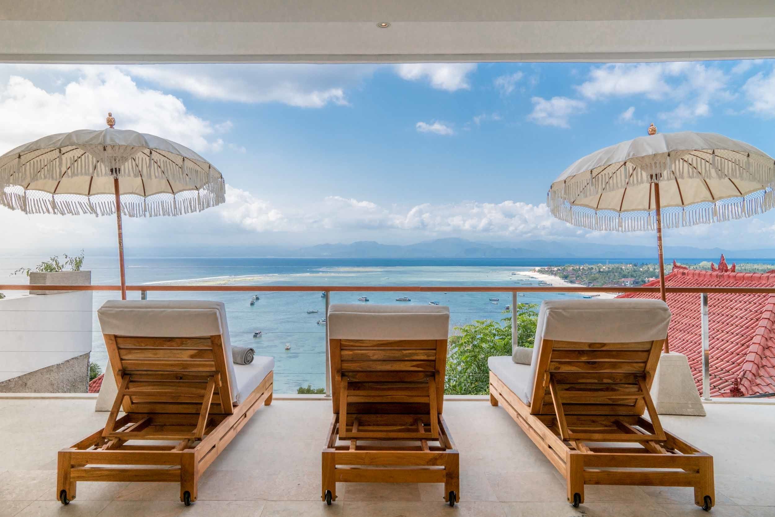 A balcony with three wooden lounge chairs and two large umbrellas, overlooking a coastal view with the sea and distant mountains.