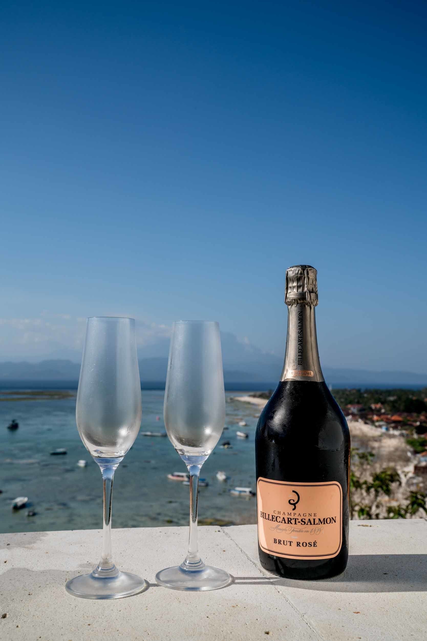 Two empty wine glasses and a bottle of Billecart-Salmon Brut Rosé champagne on a ledge with a coastal view.