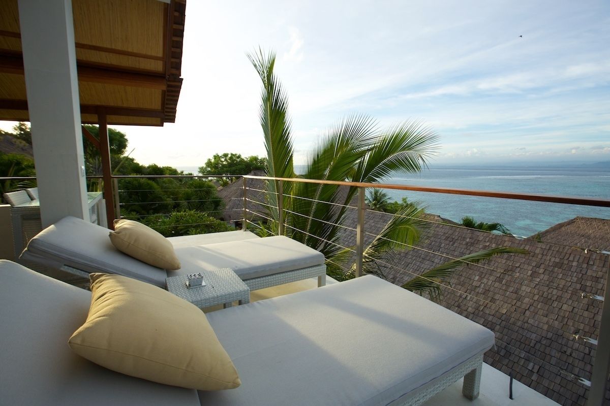 Un balcón con dos sillones de relax, uno con una almohada color beige. El balcón tiene vistas a una zona costera con palmeras y vistas al mar.