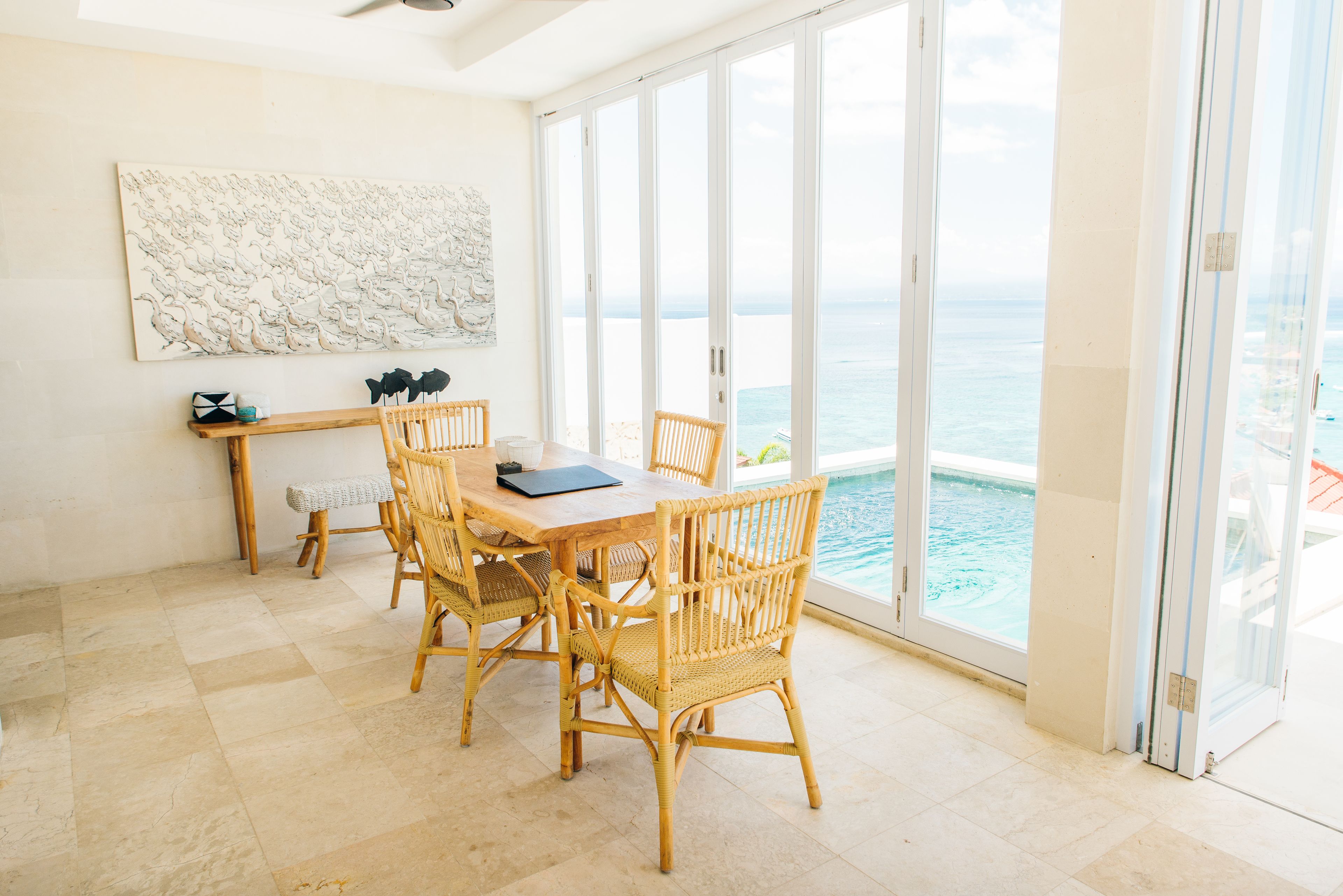A dining area with a table and chairs, a large artwork on the wall, and a view of the ocean through large sliding doors.