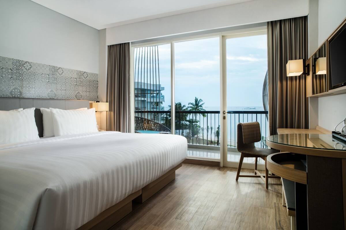 Habitación de hotel con una cama grande, un escritorio, una silla y un balcón con vista al océano y edificios.