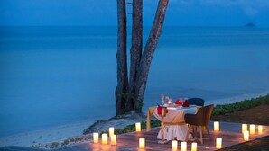 Ein romantisches Abendessen am Strand mit einem für zwei Personen gedecktem Tisch, umgeben von Kerzen, unter Palmen.