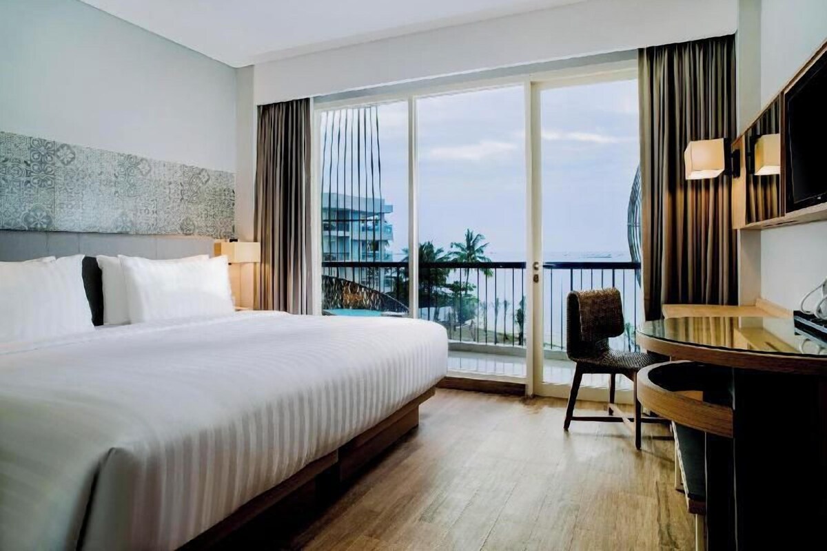 A hotel room with a large bed, a desk, a chair, a TV, and a balcony with a view.