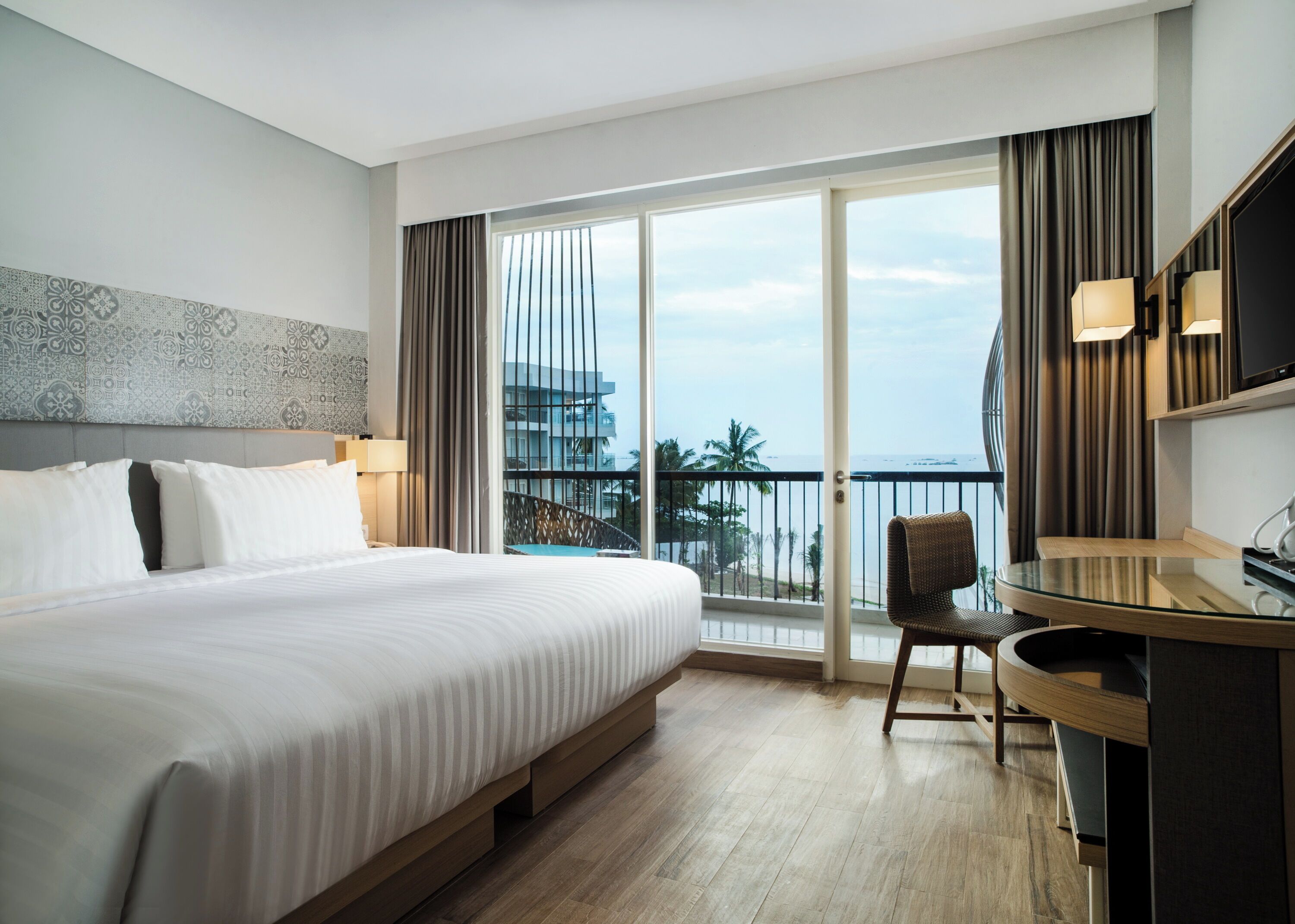 A hotel room with a large bed, a desk, a chair, a TV, and a balcony with a view of the ocean.