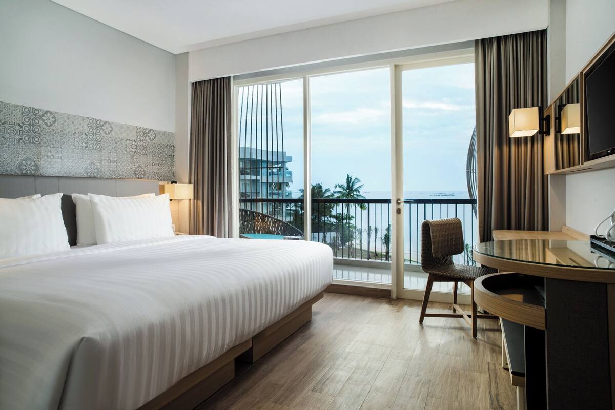 Habitación de hotel con una cama grande, un escritorio, una silla y un balcón con vista al océano y edificios.