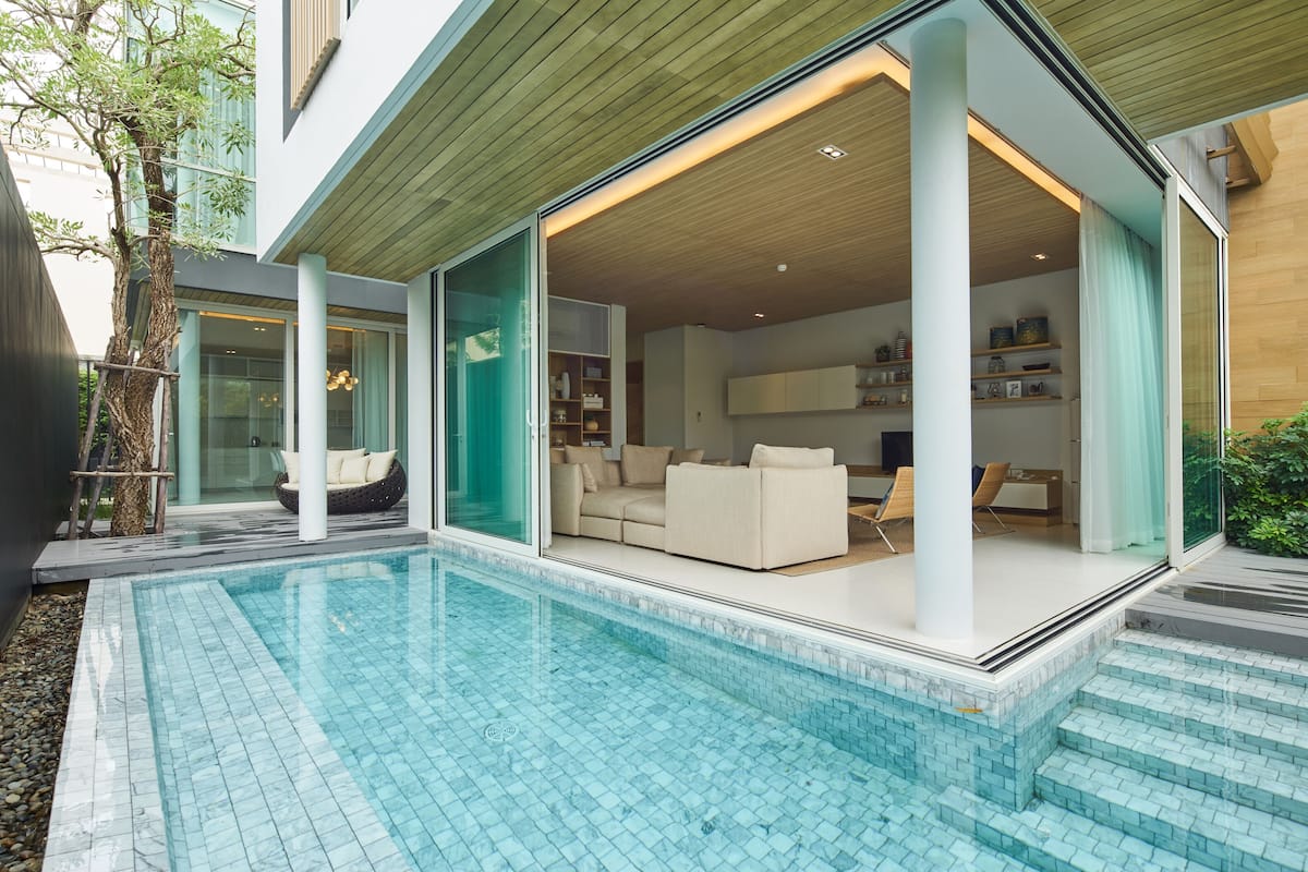 A modern house with a swimming pool, a glass door leading to a living room with a sofa and a TV, and a wooden ceiling.