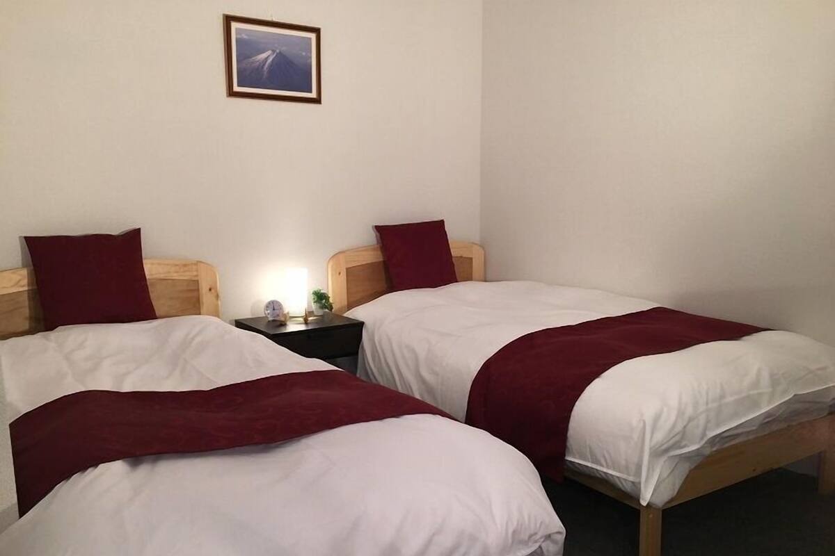 A hotel room with two single beds, each with a wooden headboard, white bedding, and a red throw blanket. There is a nightstand with a lamp and a small clock between the beds. A framed picture hangs on the wall above the beds.