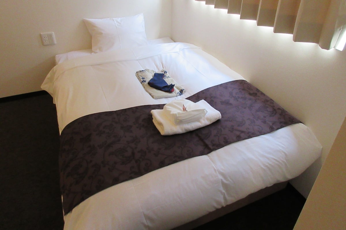A neatly made bed with a white and brown bedspread, a folded towel, and a neatly folded blue garment on the bed.