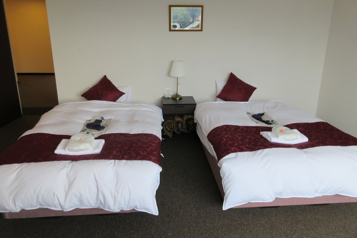 Two beds with white and red bedding, a small table with a lamp, and a framed picture on the wall.