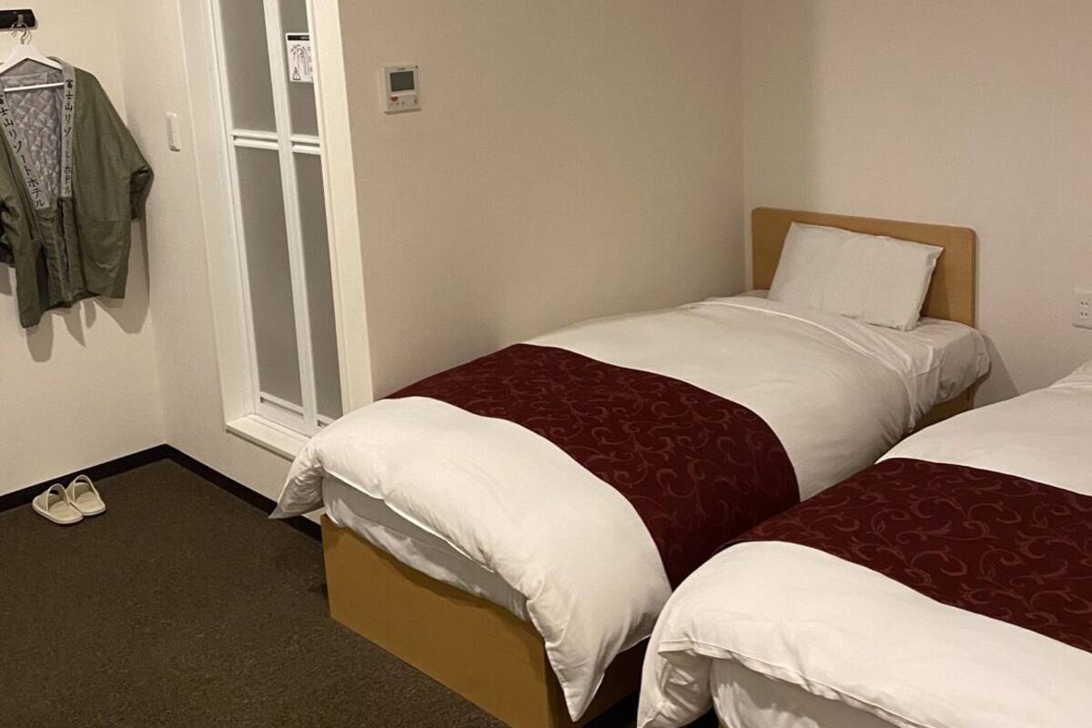 A hotel room with two beds, a window, an air conditioning unit, a coat rack, and a pair of shoes on the floor.