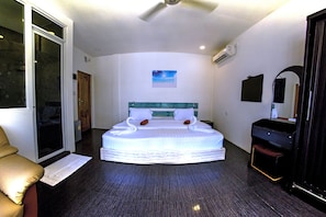 A hotel room with a large bed, a ceiling fan, a small painting, and a door leading to another room.