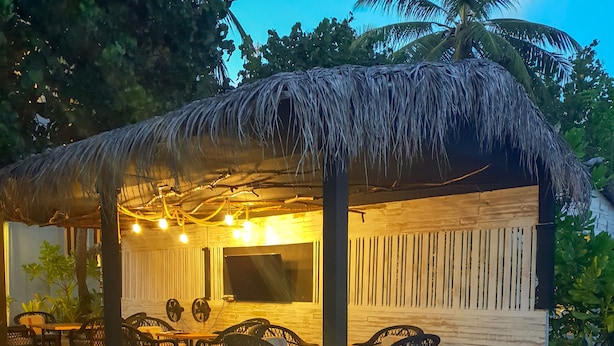 Un'area salotto esterna con tetto di paglia, tavoli e sedie in legno, un televisore a schermo piatto e luci decorative.
