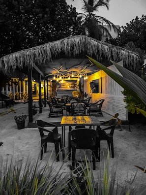 An outdoor seating area with thatched roof, tables, and chairs, surrounded by potted plants and palm trees.