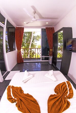 A hotel room with a bed, a ceiling fan, a TV, and a balcony with a view of trees.