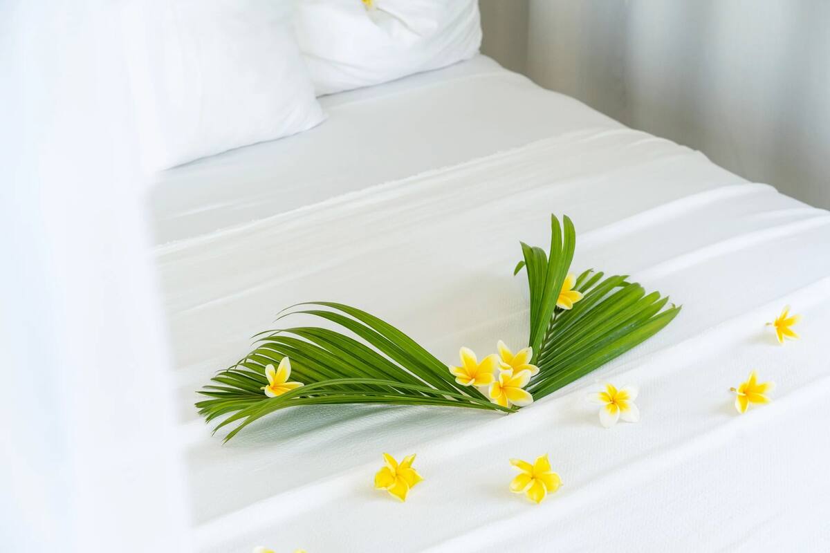 Een netjes opgemaakt bed met witte lakens, een bloemstuk met gele bloemen en groene bladeren, en een wit kussen.