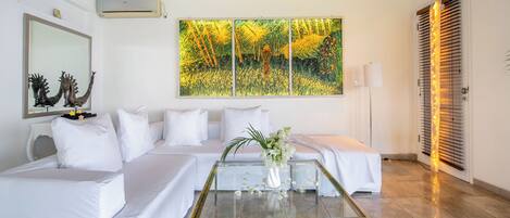 Una habitación de hotel moderna con un sofá blanco, una mesa de centro de vidrio con un arreglo floral y un cuadro de paisaje grande en la pared.