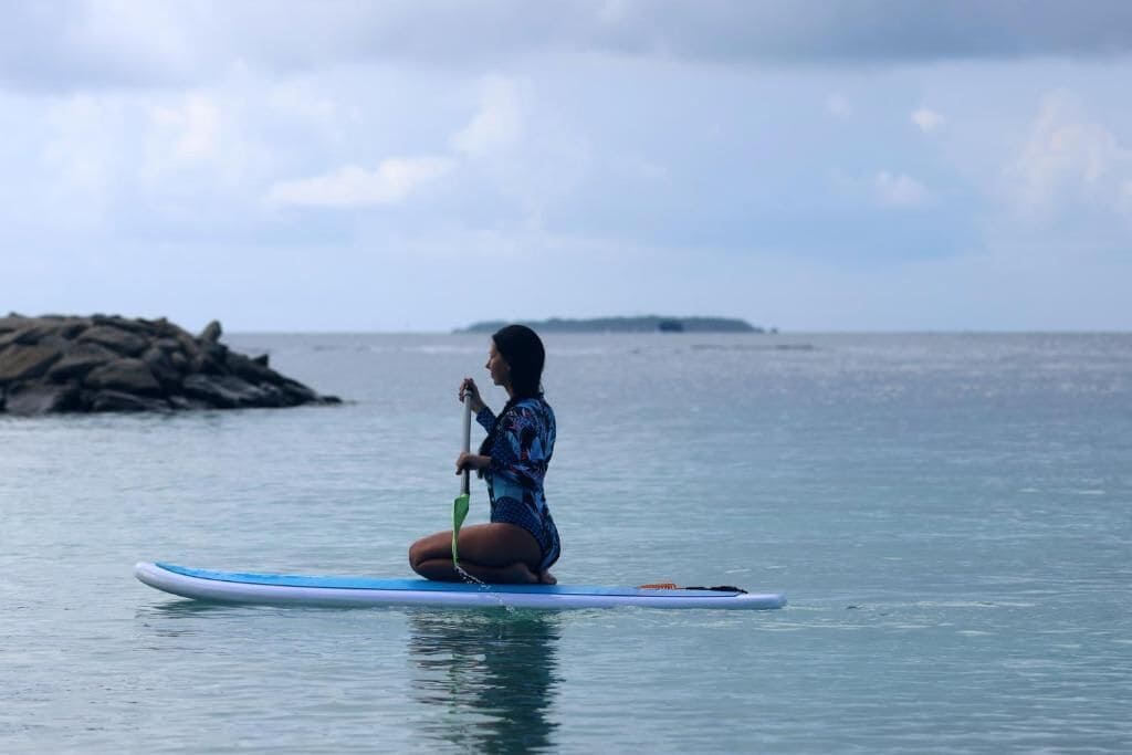 A person sitting on a paddleboard in calm waters with a rocky shoreline and distant land in the background.