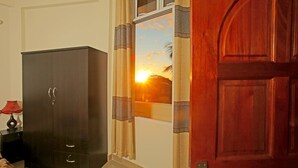 A room with a dark wooden wardrobe, a red lamp on a side table, and a view of a sunset through a window with patterned curtains.