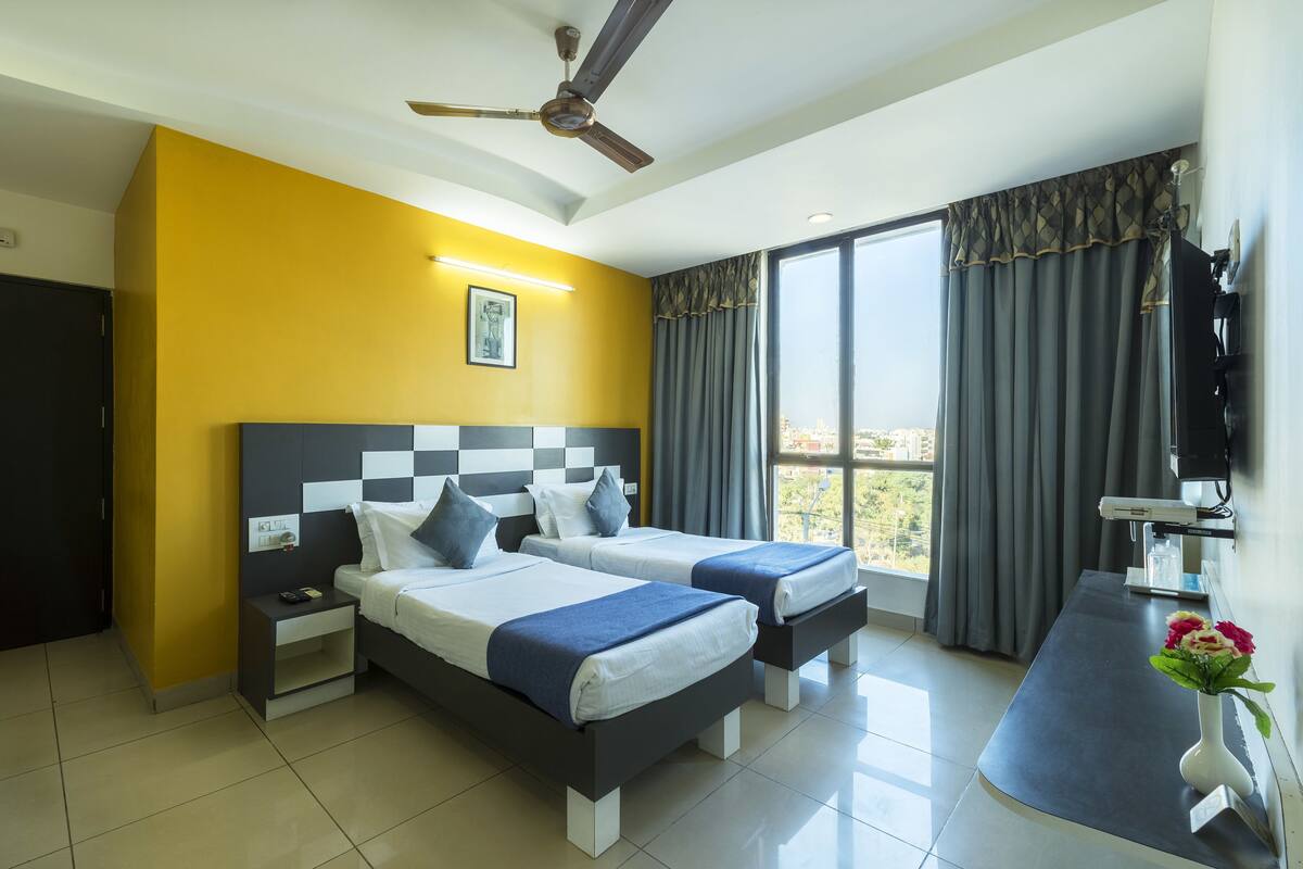 Habitación de hotel con dos camas, ventilador de techo, ventana grande con cortinas, escritorio con silla y televisor en la pared.