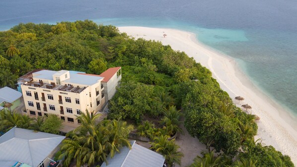 An aerial view of a resort with a beach, dense tropical vegetation, and a multi-story building with balconies.