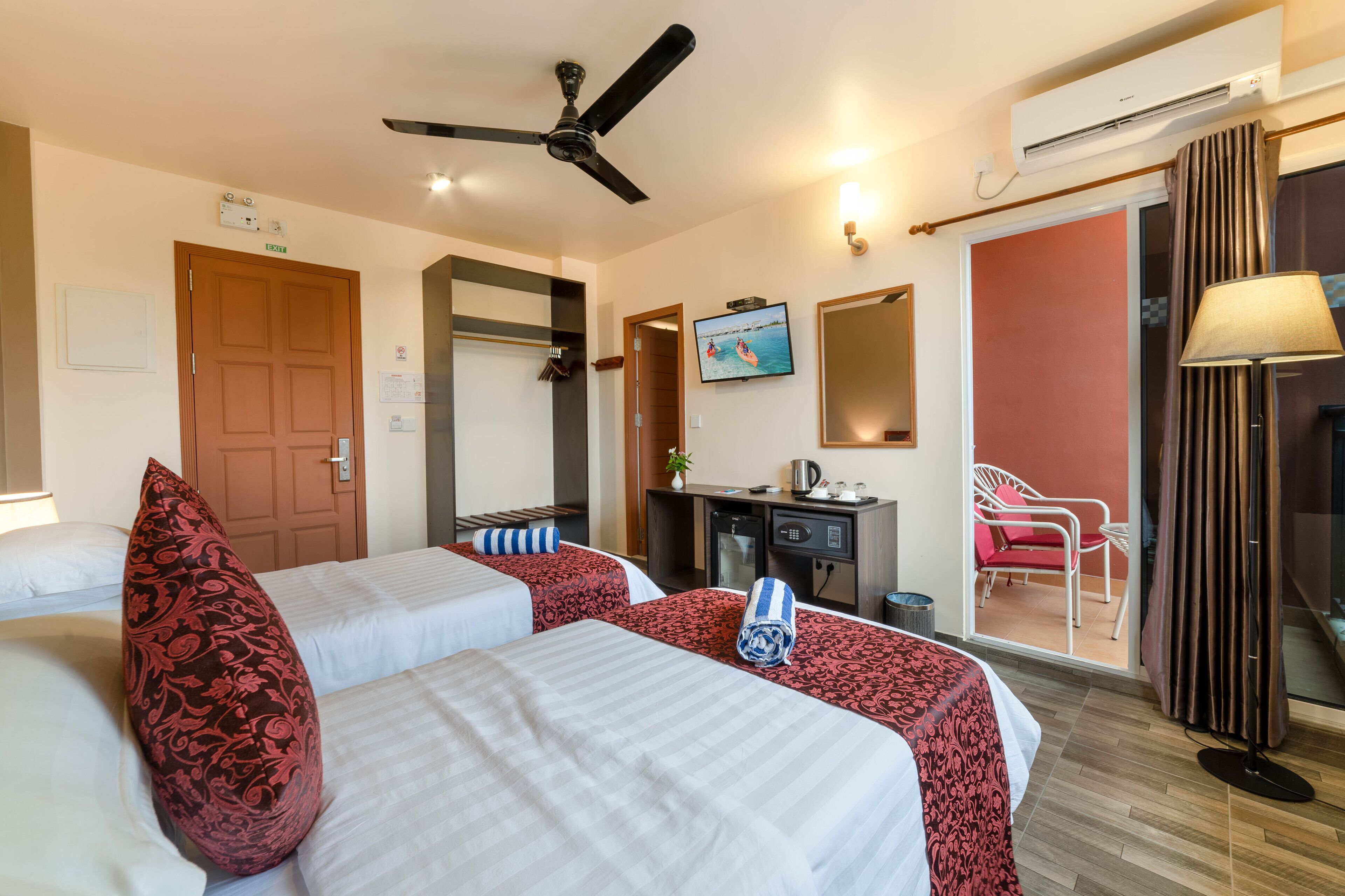 A hotel room with two beds, a ceiling fan, a TV, a desk, a chair, and an air conditioning unit.
