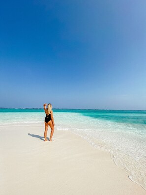 Eine Person steht auf einem Sandstrand mit klarem türkisblauem Wasser und wolkenlosem Himmel.