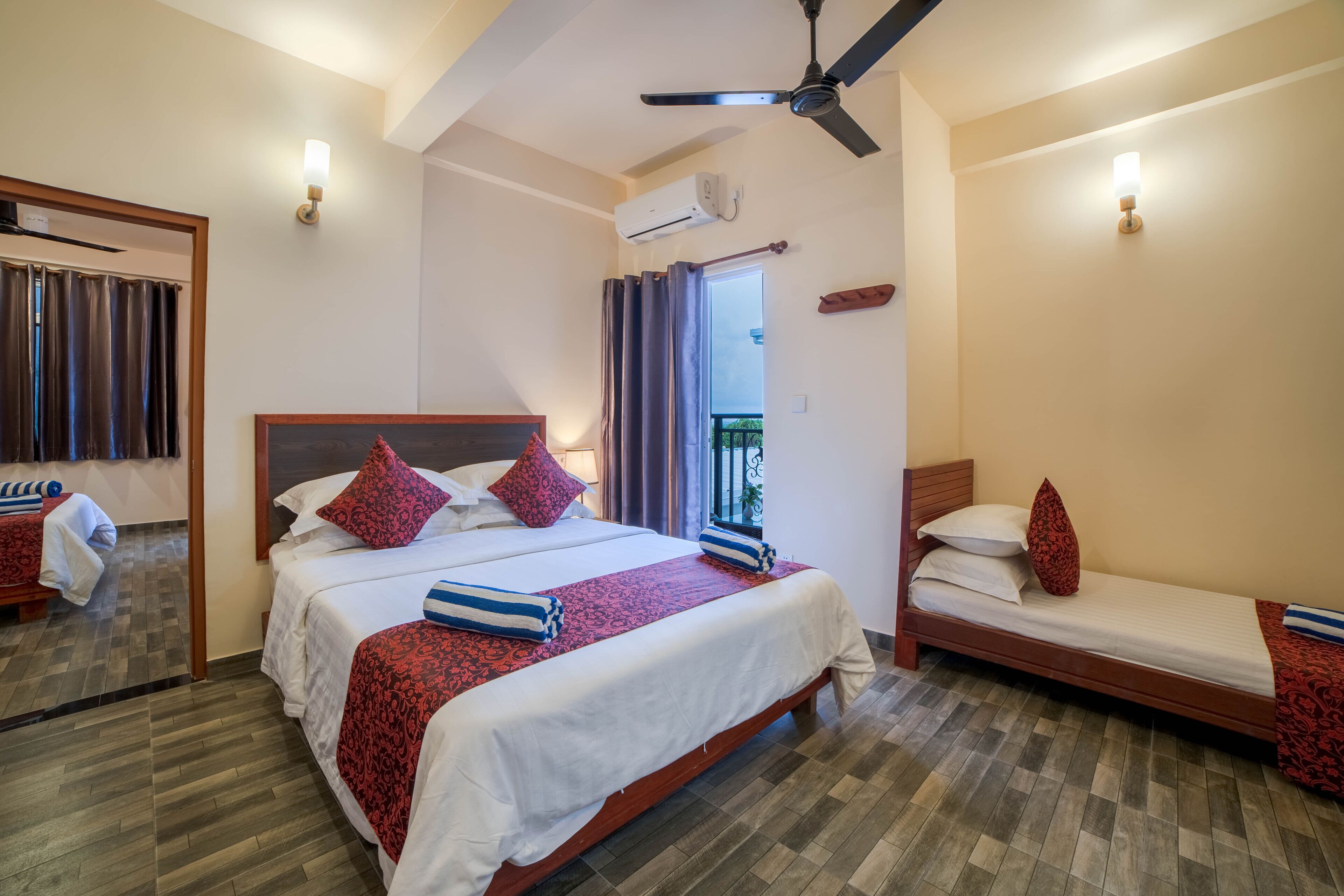 Ein Hotelzimmer mit einem großen Bett, einem kleineren Bett, einem Deckenventilator und einem Balkon mit Vorhängen.