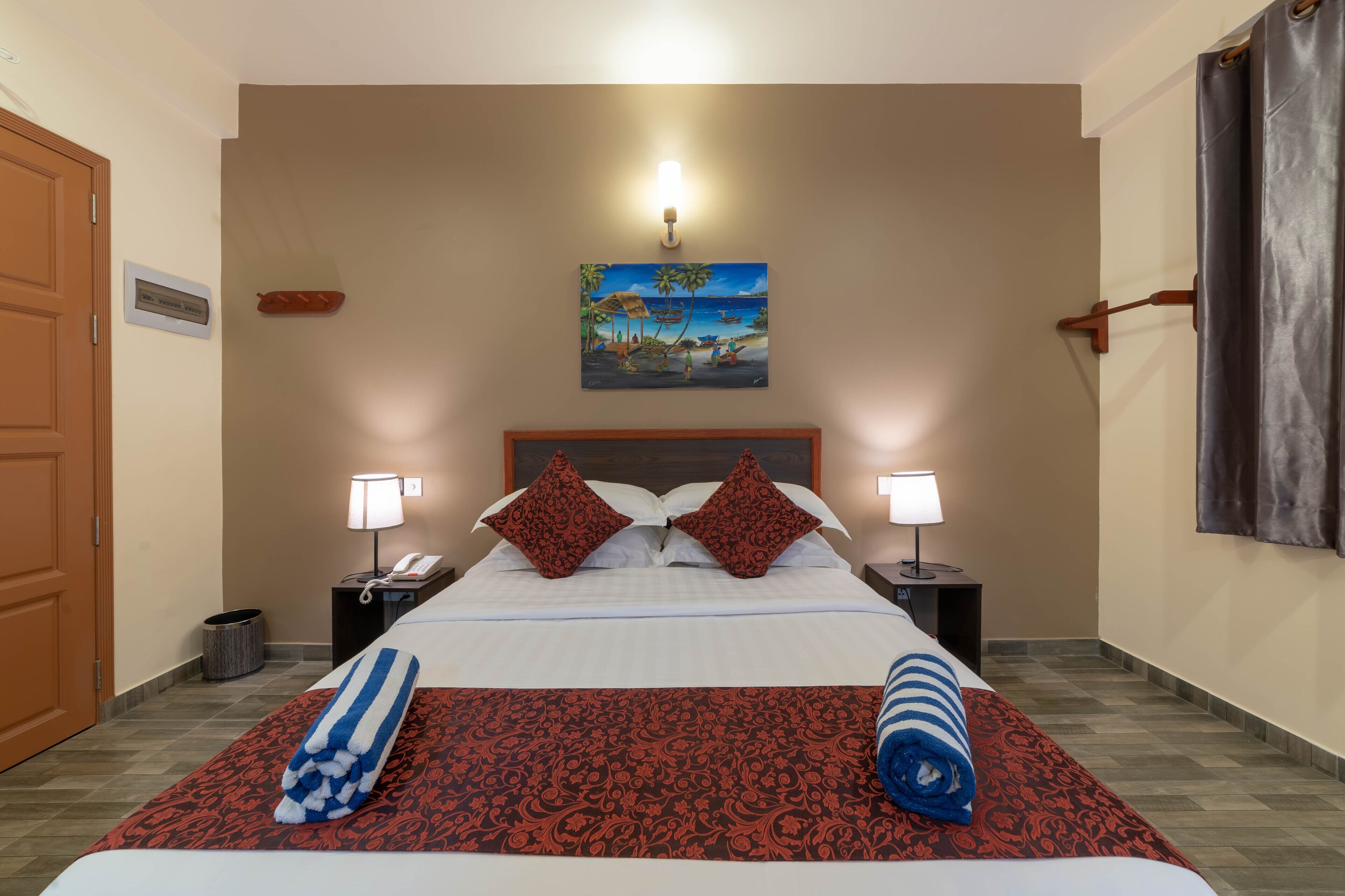 A hotel room with a bed, two bedside lamps, a painting of a beach scene, and a window with curtains.