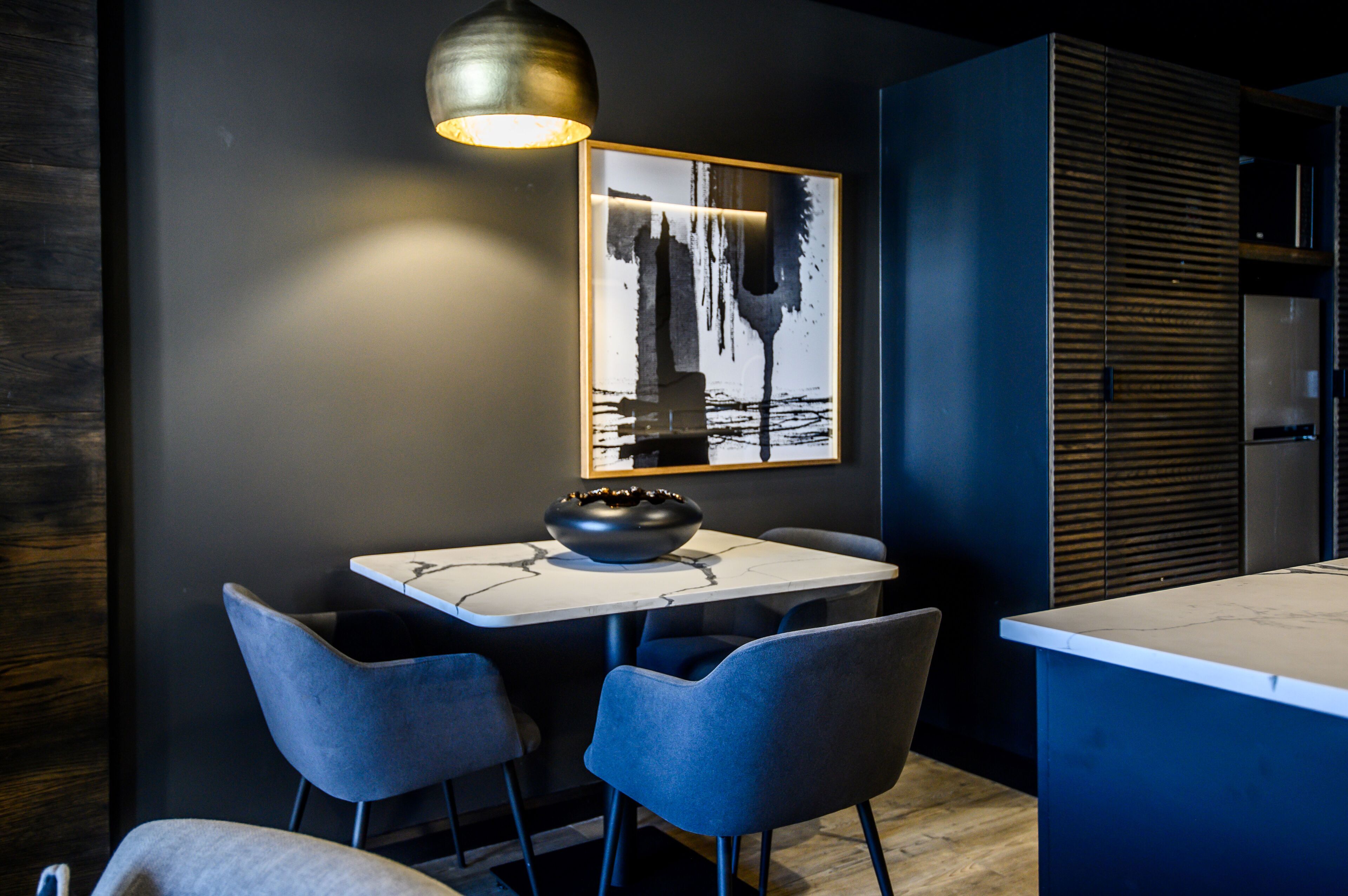 A modern kitchen with a marble countertop, two blue chairs, a black bowl, and a framed abstract painting on the wall.
