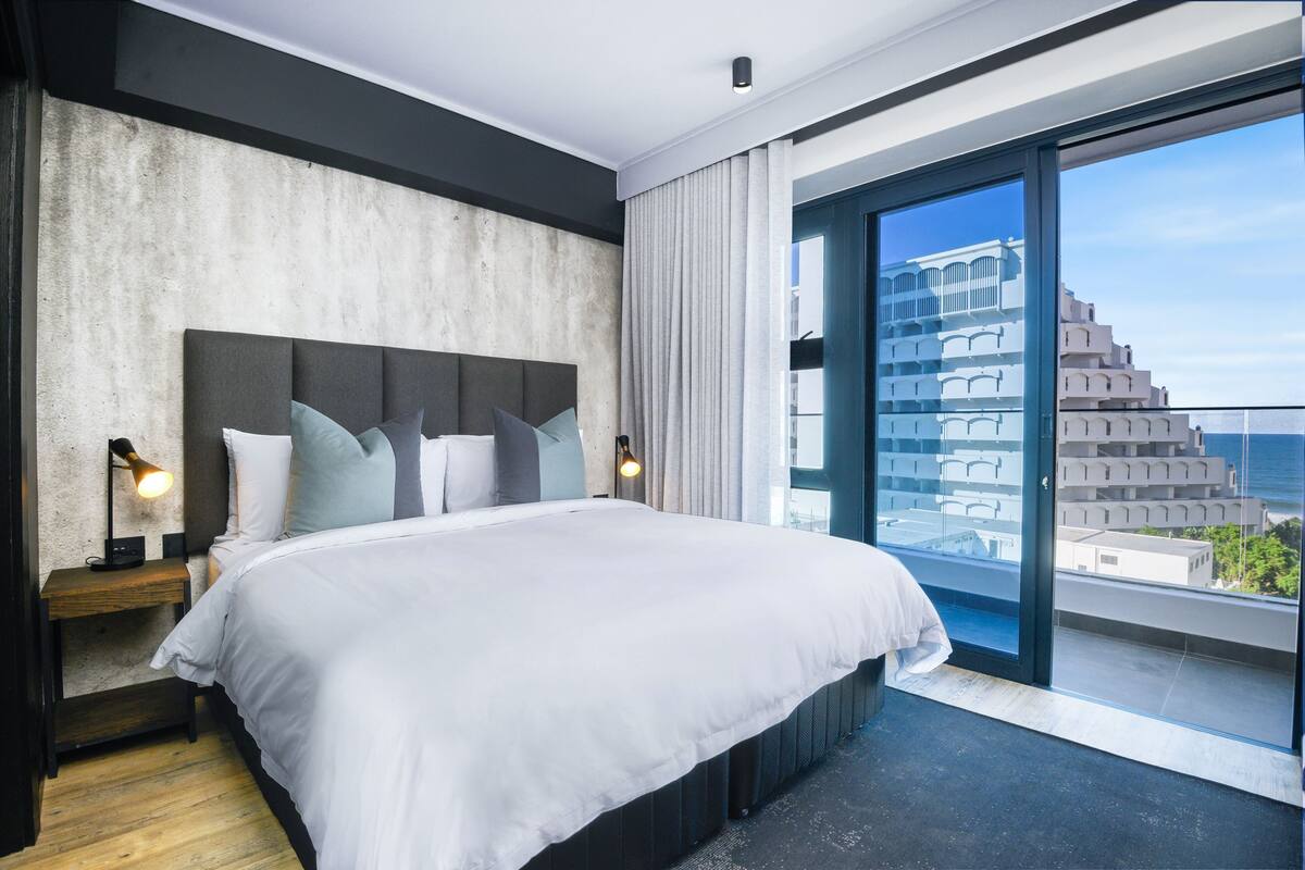 A modern hotel room with a large bed, a nightstand with a lamp, and a view of a building through a glass door.