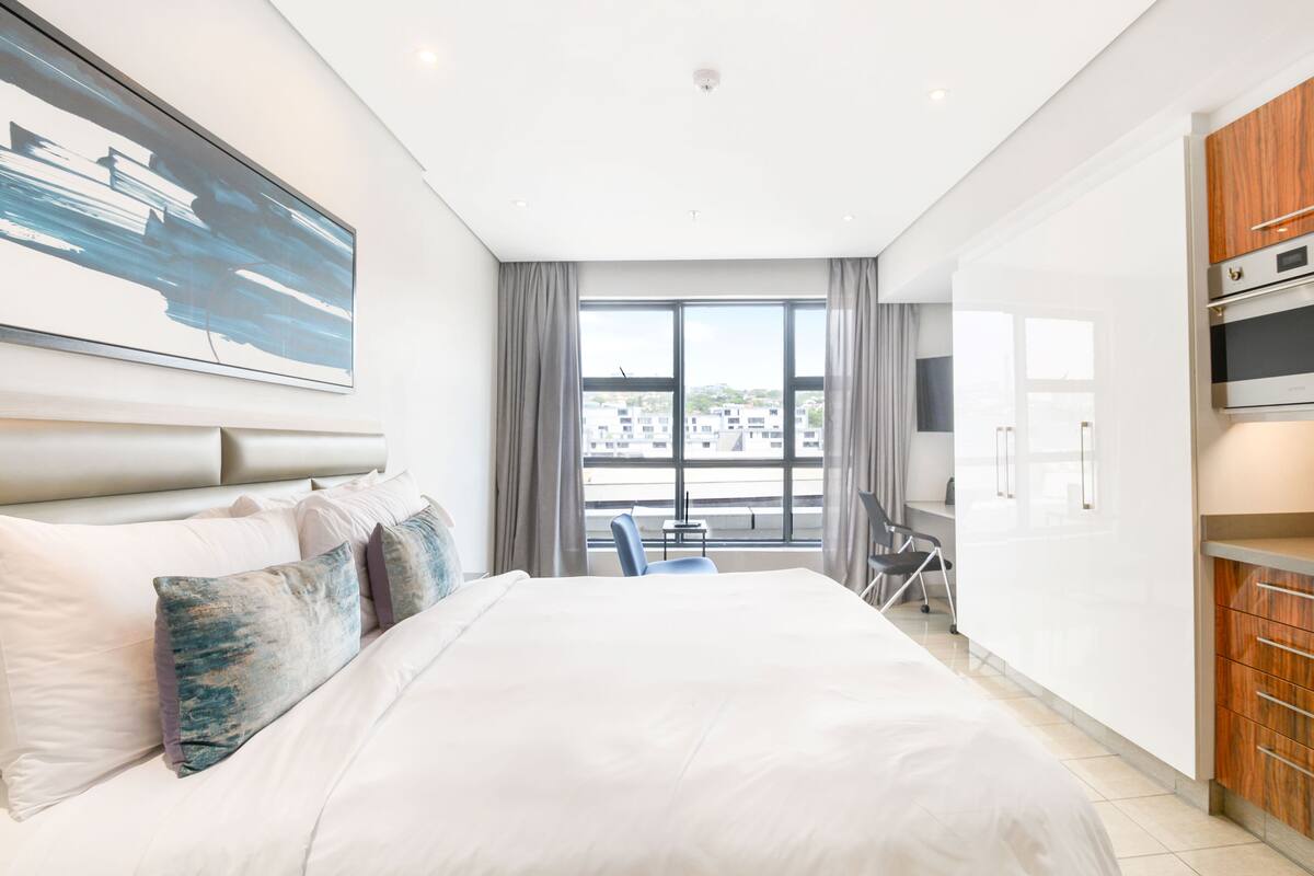 A modern hotel room with a large bed, a desk, and a view of the city through the window.