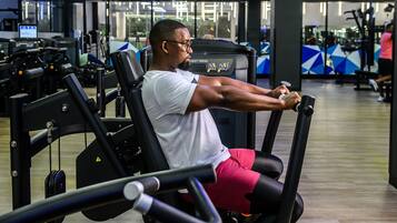 Un hombre usando una máquina de press de pecho en un gimnasio con diversos equipos de ejercicio y piso de madera.