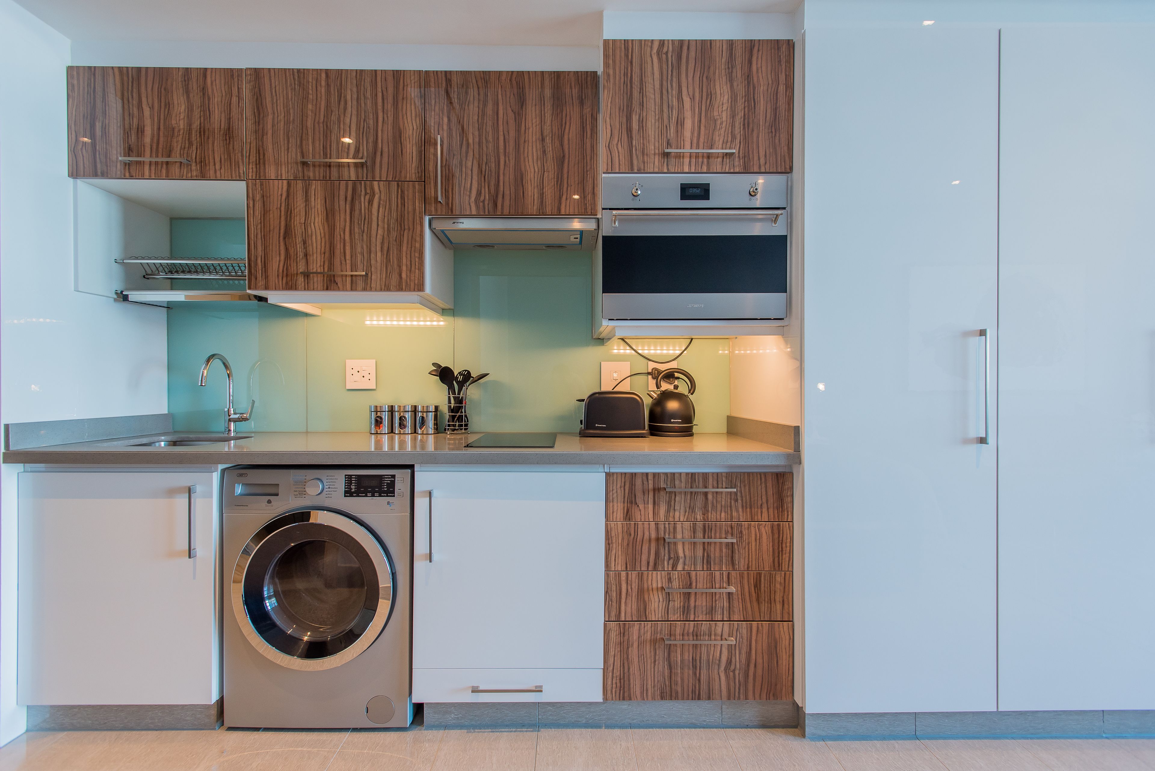 Une cuisine moderne dotée d’un lave-linge et d’un sèche-linge intégrés, de meubles en bois et d’appareils électroménagers en acier inoxydable.
