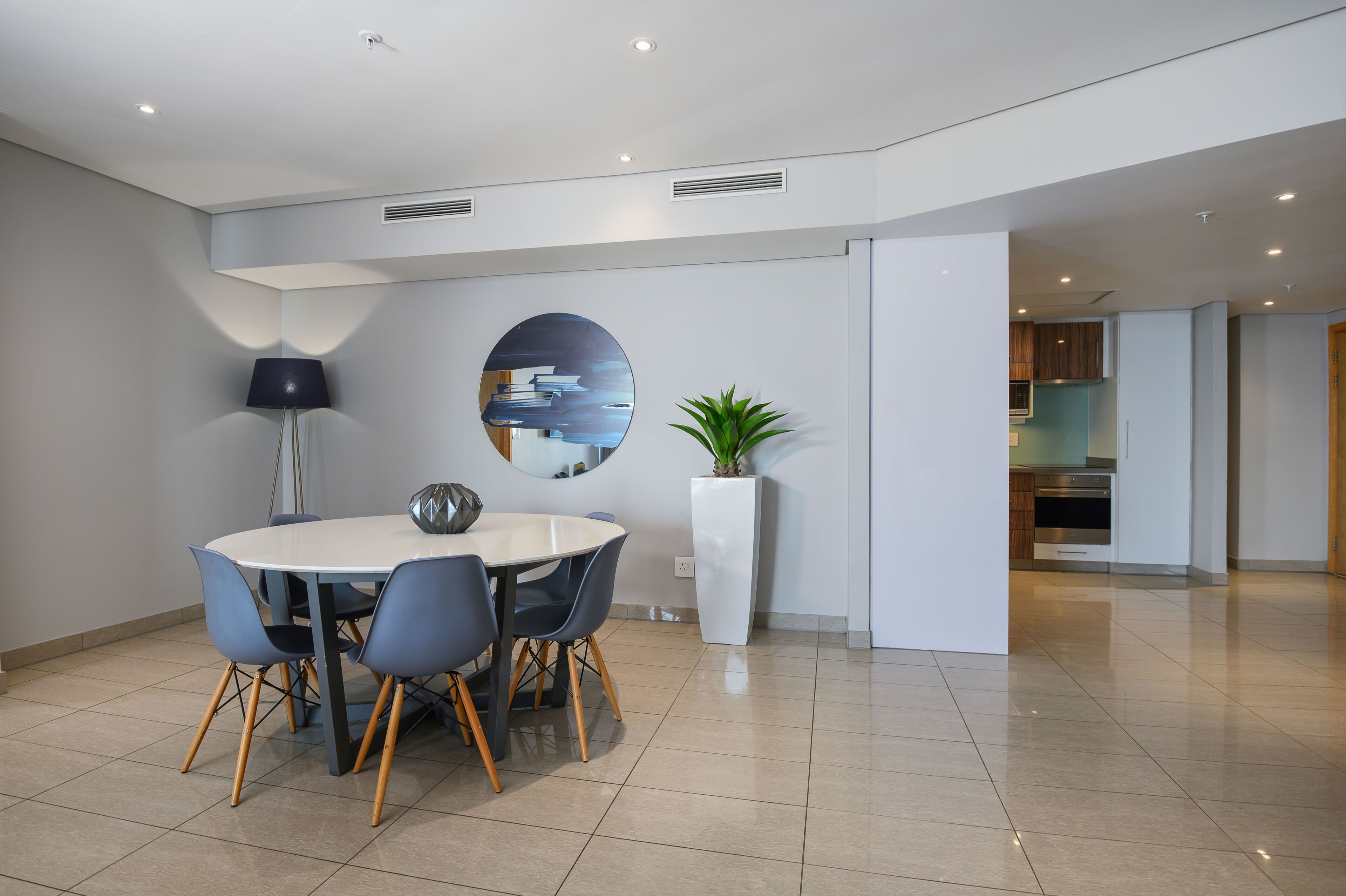 A modern dining area with a round table, blue chairs, a floor lamp, a large mirror, and a potted plant.