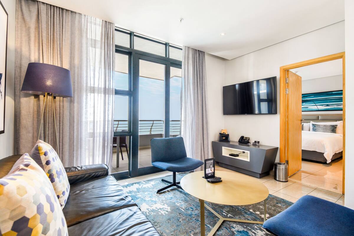 A modern hotel room with a sofa, a round table, a chair, a TV, a bed, and a balcony view.