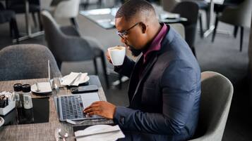 A man in a suit sitting at a table with a laptop and a cup of coffee, in a modern office setting with chairs and tables.