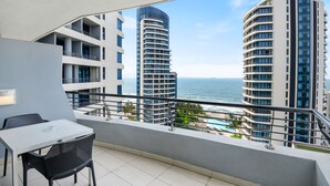 Un balcon avec une table et des chaises, donnant sur une piscine et sur deux immeubles imposants, avec vue sur l’océan.