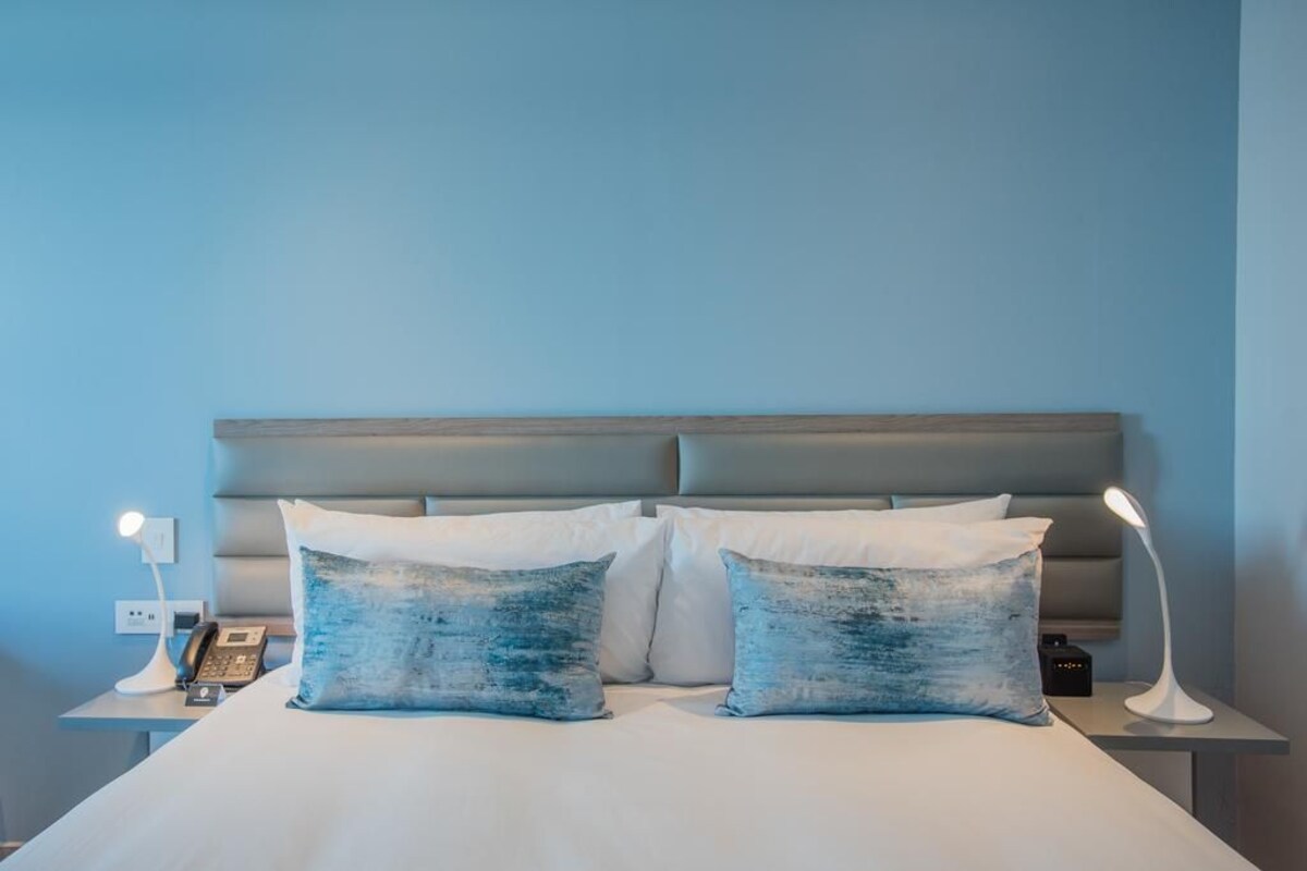 A hotel room with a bed, two pillows, a bedside lamp, a phone, and a wall socket.