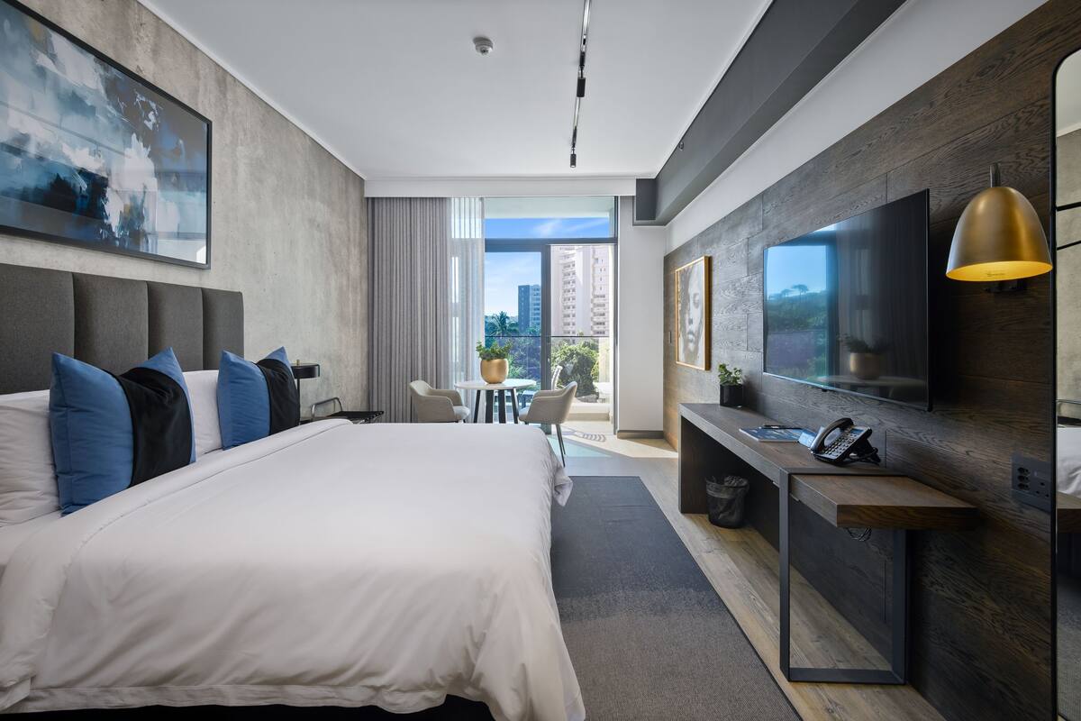 A modern hotel room with a large bed, a desk with a telephone, a flat-screen TV, and a view of the cityscape.