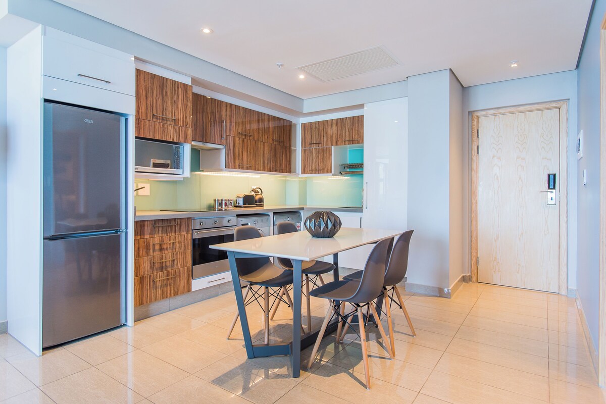 A modern kitchen with a built-in oven, microwave, and refrigerator. There is a dining table with four chairs and a decorative vase.