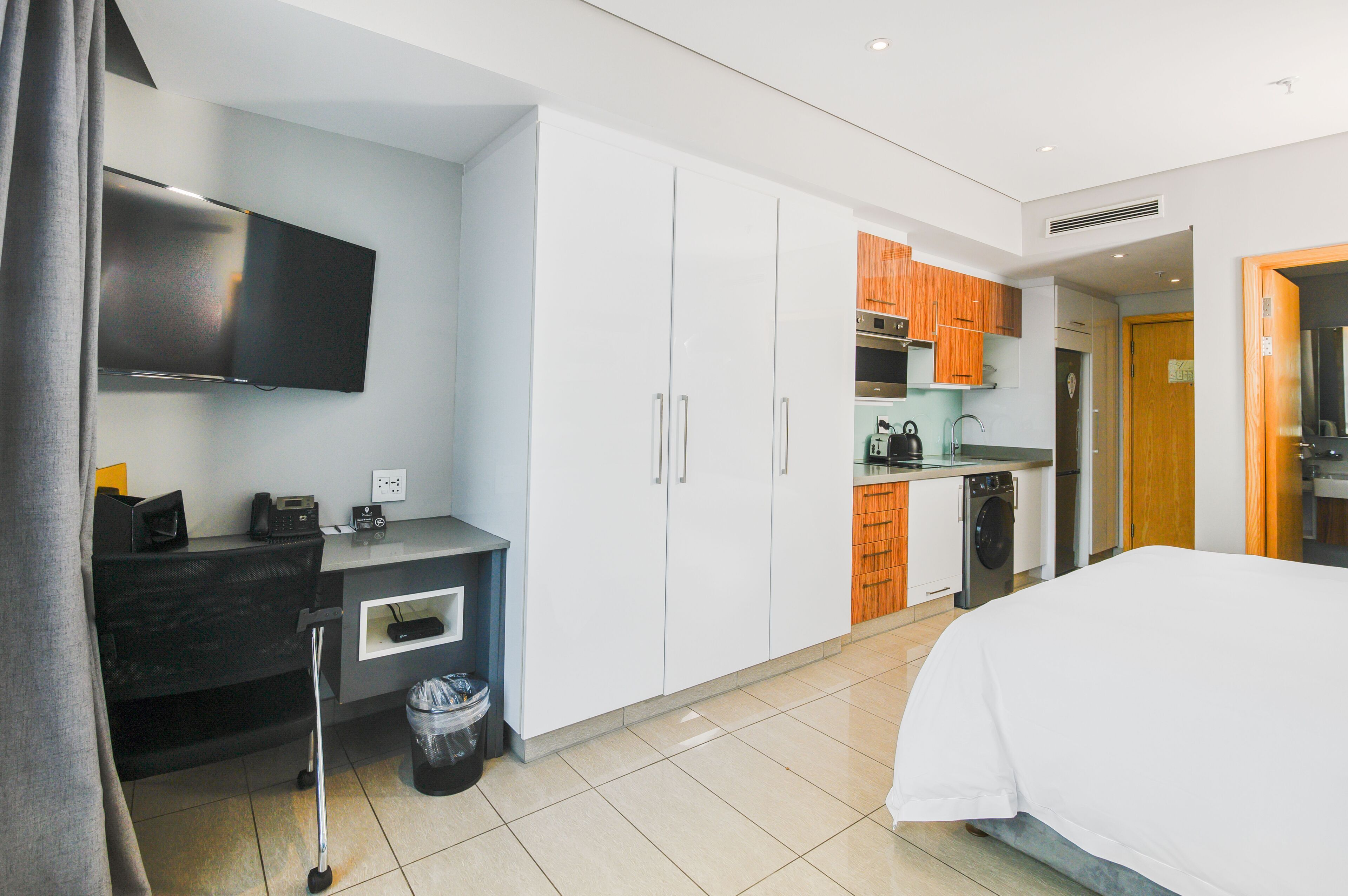 Una habitación de hotel moderna con una cama, un escritorio con una computadora, un televisor y una cocina equipada.