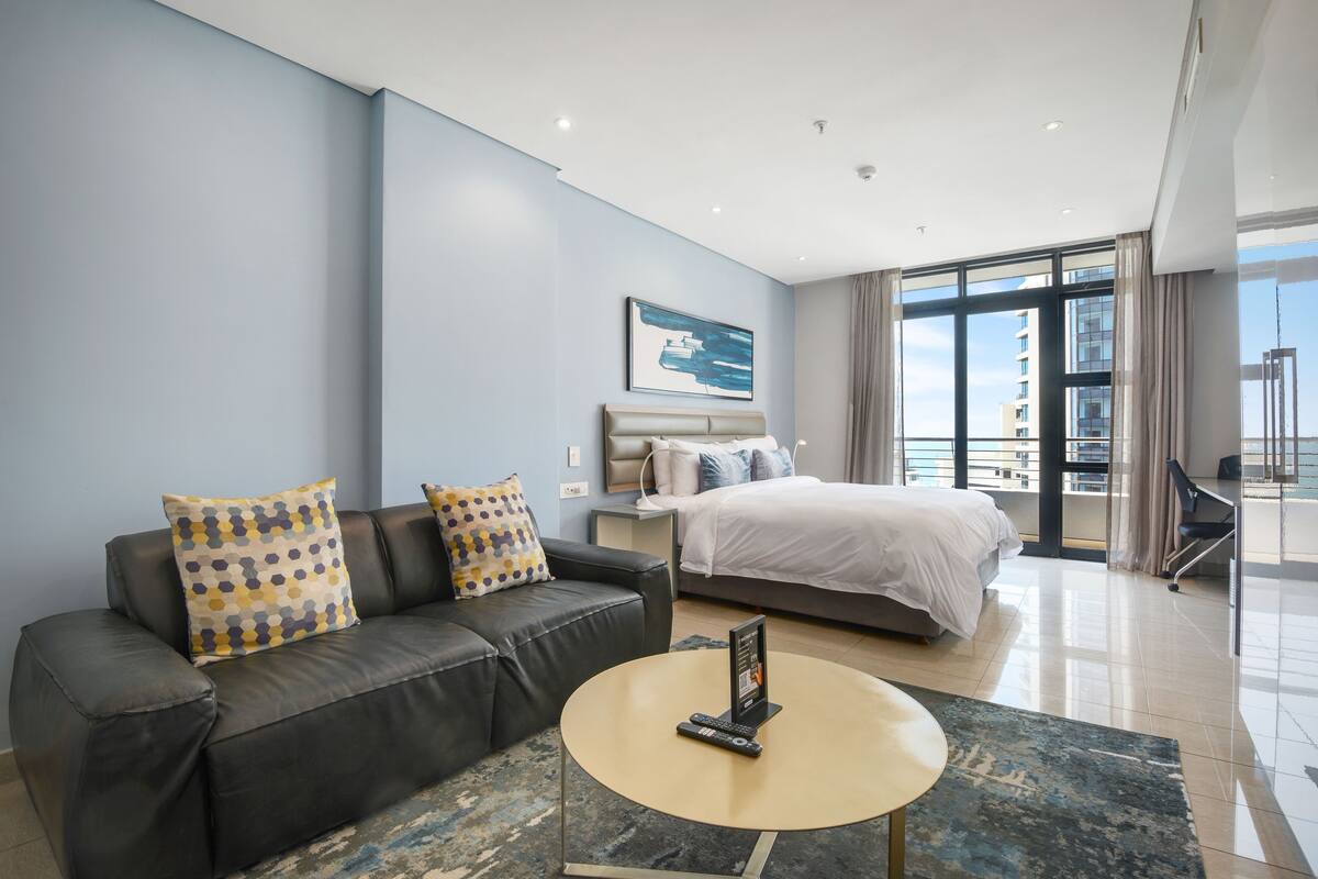 A modern hotel room with a grey leather sofa, a round coffee table, a bed with a white headboard, and a large window offering a city view.